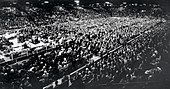 Thousands of spectators in a large arena look towards a man standing behind a podium on a platform