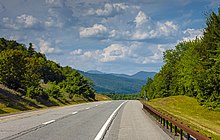 A two-lane roadway with a white dashed line in the center, white solid line at the right edge, dashed yellow line at the left and brown metal guardrail alongside the left edge, with woods on either side, drops down a slope in the foreground. In the distance, blue with haze, is an irregular, ragged ridgeline