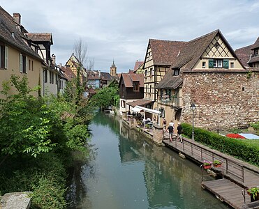 Maisons à colombages au bord d'une rivière. À droite, un quai ; à gauche des arbres ; en arrière plan, on aperçoit un clocher.