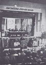 Man speaking at a podium in front of six people at a dais and a seated audience