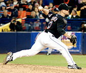 A left-handed man in his mid-thirties wearing a black baseball jersey and cap and white baseball pants throws a baseball from a pitcher's mound.