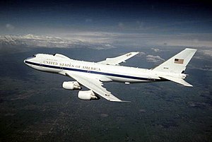 White jet with blue cheatline in-flight over land, flying left. The aircraft features a distinctive bump above the usual 747's trademark nose profile