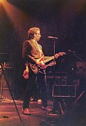 A man dressed in a black suit holds a guitar while singing into a microphone.