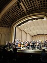 In a ivory concert hall, a stage full of classical musicians and conductor are standing at the end of a performance