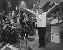 Black and white photograph from the 1958 contest held in the AVRO Studios in Hilversum, the Netherlands; an orchestra seated to the left of a small stage, with Italian singer Domenico Modugno singing on the stage platform in front of a wall.