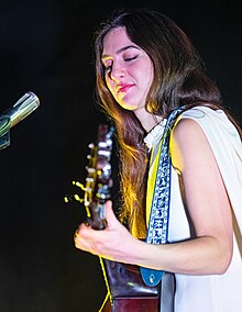 Stagelights illuminate a female singer-songwriter against a black background as she performs with her guitar (hanging from a strap) and smiles