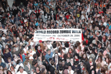 A packed crowd in zombie makeup hold a banner reading, "World Record Zombie Walk, Pittsburgh, Pennsylvania, October 26, 2006, The It's Alive Show, Pittsburgh East Nissan, Monroeville Mall"