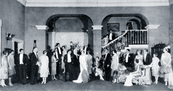 stage, with set depicting a ballroom full of guests in evening dress; centre is a woman in a white gown