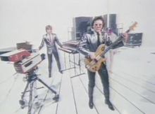 In a white studio, Geoff Downes is playing multiple keyboards and Trevor Horn playing a bass guitar, both wearing silver suits. A woman in a tube behind Horn is also wearing a silver costume.