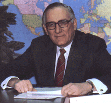 Middle-aged man with black and gray hair sitting behind a desk, wearing a navy blue suitcoat, white dress shirt, red necktie, and black-rimmed eyeglasses