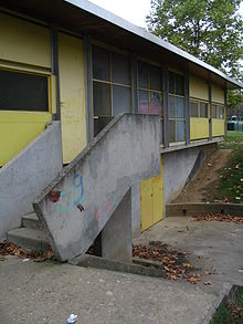 Photographie en couleurs représentant un bâtiment préfabriqué avec un escalier d’accès en béton au premier plan.