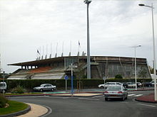 Photographie du stade de béton brut avec à gauche la tribune couverte et à droite un mur courbe fait de baies vitrées renforcées par des structures en V. Par-dessus le toit plat surgissent une douzaine de piliers de béton terminés par des mâts.