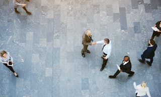 From above two adult business men shaking hands met in office hall with workers walking around.