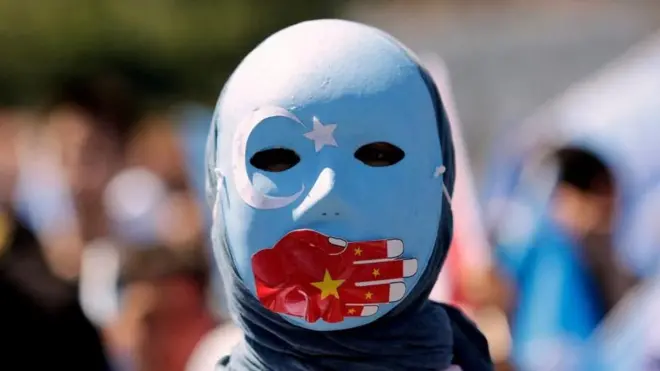 一名维吾尔族抗议者（他戴着饰有星月符号的蓝色面具，一只红手举着中国国旗捂住嘴）