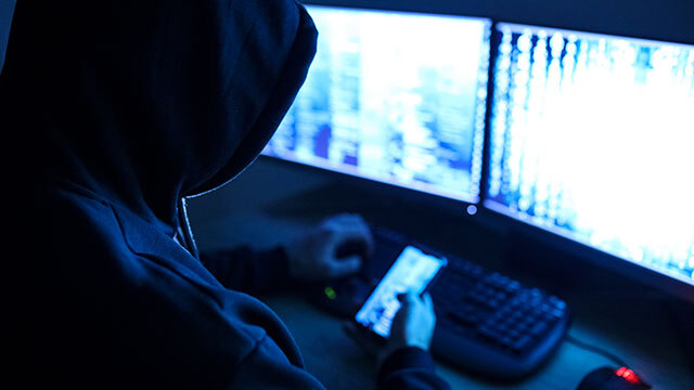 Hooded man using mobile phone in front of a computer screen to commit fraud and scams