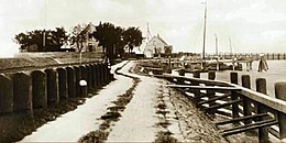 Photographie en noir et blanc d'un chemin longé par une digue et un port au premier plan et de maisons en second plan.