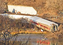 An area of land with brown vegetation lit by golden late-afternoon sunlight next to water and railroad tracks in the background at the base of a steep hill with a white concrete retaining wall. A silvery rail car lies along the shore, almost all the way to the water, with its windows either broken out or severely cracked. Behind it is another car, lying almost on its side.