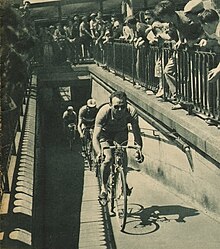 Un homme avec des lunettes de soleil sur un vélo devance deux autres cyclistes, entouré par une foule qui les observe.