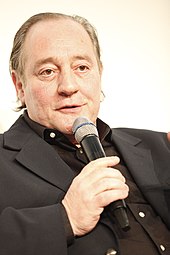Portrait d'un homme habillé avec une chemise et une veste noire. Il tient un micro avec sa main droite et semble en train de s'exprimer.
