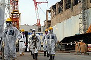 Plusieurs personnes en combinaison devant un bâtiment partiellement détruit.