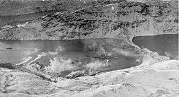 Black and white photograph of a World War II-era warship in a small bay with a steep and snow-covered shoreline. Clouds of smoke are being blown from the shoreline towards the ship.