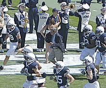 Un vieil homme en costume court entre des joueurs de football américain en tenue, devant un orchestre universitaire qui joue.