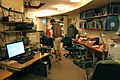 An interior view of a curved, windowless room with desks along the sides. On the desks are computers and electronic instruments. In front of the desks are an oxygen bottle and an oscilloscope sitting next to an empty oscilloscope cart. Above the desks are shelves mounted to the walls, which hold more electronic instruments and a collection of binders. In the middle distance, three people (two seated and one standing) look at a computer screen which is hidden from our view by the curve of the room. In the background is an old Polycom video conferencing system with a large CRT TV on a cart, next to a bookshelf with magazine boxes, hard hats, and other items.