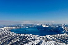 Blick von einem Berg auf den gesamten Caldera-See, mit schneebedeckter Umgebung bei blauem Himmel.