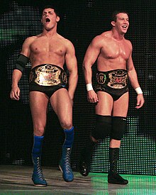 Two dark-haired men are both wearing black wrestling tights. One wears knee-high blue wrestling boots with blue kneepads and a black elbow pad on his right arm, and the other wears black knee-high wrestling boots with black kneepads, and white tape around his wrists. The two are wearing professional wrestling championship belts around their waists.