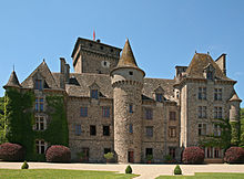 Sur cette photo, à l'arrière-plan sous un ciel d'un bleu pur, le donjon du château. D'une hauteur de 40 mètres, en forme de lourd parallélépipède un peu élargi tout en haut, il date des débuts de la guerre de cent ans et domine le corps de logis, datant du XVIIe, mais transformé au XIXe selon la mode troubadour. L'aile gauche est une construction qui en fait partie, mais en ressort un peu avec son toit pyramidal avec, à l'extrême gauche, une tour ronde au toit pointu, moins élevée. À la droite du corps de logis, une construction plus détachéede l'ensemble tout en y demeurant liée aussi, plus haute, surmontée d'un toit pyramidal tronqué à quatre faces. Au centre une haute et plus grosse tour, plus détachée du reste, également au toit pointu, dépasse tout et même (mais par illusion d'optique), le donjon médiéval.