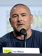 A middle-aged white man in a T-shirt, speaking into a microphone.