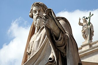 Untersicht in Farbfotografie von einer männlichen Statue mit Stola. Er hält eine aufgerollte Schriftrolle auf seinem linken Unterarm. Im Hintergrund ist auf einem Gesims eine männliche Figur mit einem Heiligenschein aus Eisen zu sehen. Er hält ein Eisenkreuz an seiner linken Seite und hebt seinen Arm nach oben, mit drei ausgestreckten Fingern.