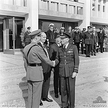 Photographie en noir et blanc représentant deux officiers se serrant les mains, devant les yeux de leurs subalternes.