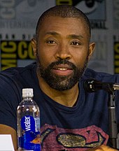 Uma fotografia de Cress Williams falando em uma convenção atrás de um microfone