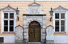 Frontale Farbfotografie vor dem Eingang einer orangen Fassade. Die Tür ist mit roten Querstreifen und goldenen Blumen verziert. Im oberen Teil ist eine Figur in einem Wappen. Um die Tür sind mit grauem Stein Ornamente eingemeißelt, die auch in einem Quadrat und einem Giebelfeld über der Tür erscheinen. Im Quadrat halten zwei Löwen ein Wappen. An beiden Seiten der Tür stehen zwei gemeißelte Steinblöcke und zwei Laternen hängen zwischen Tür und zwei Fenstern.