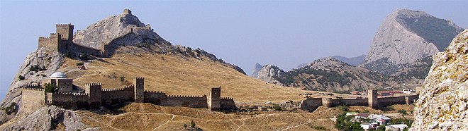 Autour d'une colline rocheuse et au sol aride une muraille de pierre crénelée et jalonnée par des tours défensives serpente jusqu'au sommet