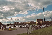 Several rows of cars parked in an outdoor parking lot under a blue sky with billowy clouds. Many buildings are in the background.