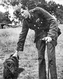 Photo noir et blanc montrant Edward Mannock debout se penchant pour carresser un chien. Il s'appuye sur une canne.