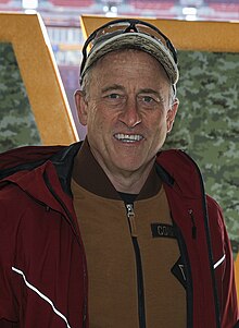 Man smiling while wearing a baseball cap and an unzipped red jacket on top of a tan sweater.