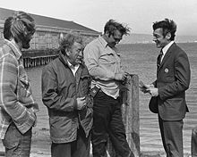 A black and white photograph of Milk in a suit with short hair speaking with three longshoremen standing by San Francisco Bay