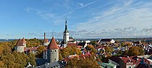 Farbfotografie in der Obersicht von einer Kirche, die von alten Gebäuden und Türmen umgeben ist. Dazwischen und vor allem im linken Bereich stehen Bäume. Im Hintergrund ist eine Bucht mit Schiffen zu sehen.