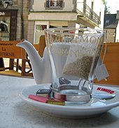 Sur une terrasse de café, gros plan sur une verre posé sur une soucoupe avec des dosettes de sucre et, au second plan, une théière blanche