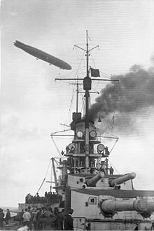 The gun turrets of a battleship. A grey dirigible flies overhead.