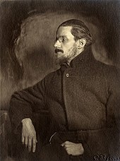 Half-length portrait of man in his thirties. He looks to his right so that his face is in profile. He has a mustache, a thin beard, and medium-length hair slicked back, and wears a pince-nez and a plain dark greatcoat, looking vaguely like a Russian revolutionary.