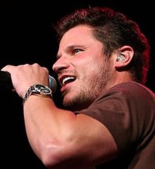 Nick Lachey singt in ein Mikrofon, das er in der linken Hand hält. Er trägt ein dunkelbraunes T-Shirt, einen Dreitagebart, einen leicht nach oben stehenden braunen Kurzhaarschnitt, einen silbernen Kopfhörer im linken Ohr und eine silberne Armbanduhr am linken Handgelenk.