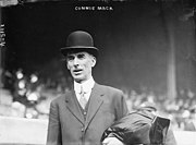 A man standing in a baseball stadium wearing a suit and bowler hat. He has a coat folded over his left arm.