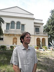 Le chercheur est photographié en contre plongée avec dans le fond la façade principale d'un bâtiment. Chemise blanche col ouvert, lunettes.