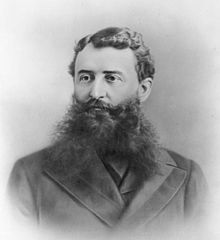 Black and white portrait of a man wearing a coat, shown from chest up, well combed and with a voluminous beard.