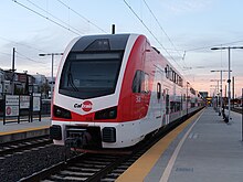 Stadler KISS 308 Caltrain von schräg vorne an Bahnsteig in Dämmerung. Links hinten Stadtvillen, davor ein anderer Bahnsteig. Im Zentrum des Bilds ist an Mittelbahnsteig lange Caltrain-Komposition, weiß lackiert mit rot aerodynamisch fließend abgesetzten Flächen und dunkelgrauen Akzenten.