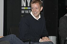 Matt Hubbard, a caucasian male with curly blond hair, wears a black sweater with a white shirt under. He smiles and looks to his right.
